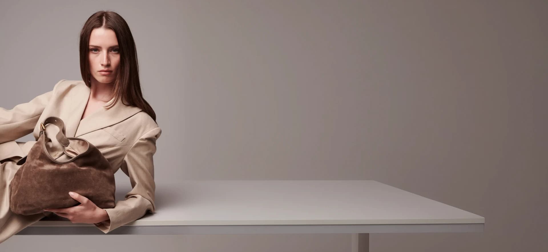 Campaign image — model reclining on a table in the beige suit with a suede bag